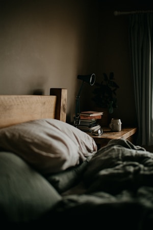 A cozy bedroom setting features a wooden headboard, soft pillows, and a neatly made bed. On the bedside table, there is a black lamp, a stack of books, and a mug. A plant adds a touch of greenery to the otherwise muted color palette. Curtains hang to the side, suggesting a window nearby.