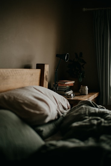 A cozy bedroom setting features a wooden headboard, soft pillows, and a neatly made bed. On the bedside table, there is a black lamp, a stack of books, and a mug. A plant adds a touch of greenery to the otherwise muted color palette. Curtains hang to the side, suggesting a window nearby.