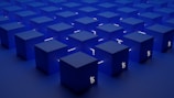 A series of identical blue cubes are arranged in a grid pattern on a flat surface. Each cube is illuminated with a subtle light, casting soft shadows. A symbol resembling the Tezos blockchain logo is visible on the sides of the cubes.