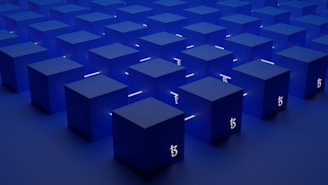 A series of identical blue cubes are arranged in a grid pattern on a flat surface. Each cube is illuminated with a subtle light, casting soft shadows. A symbol resembling the Tezos blockchain logo is visible on the sides of the cubes.