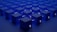 A series of identical blue cubes are arranged in a grid pattern on a flat surface. Each cube is illuminated with a subtle light, casting soft shadows. A symbol resembling the Tezos blockchain logo is visible on the sides of the cubes.