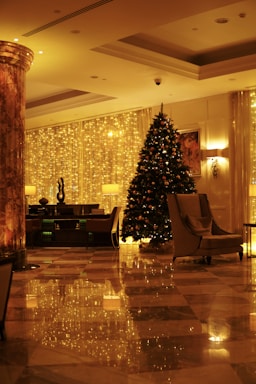 A luxurious indoor space decorated for the holiday season. A large Christmas tree adorned with lights and ornaments stands near a curtain of golden fairy lights. Elegant furniture, including armchairs and a table with a lamp, are arranged neatly. The marble flooring reflects the warm, festive lighting.