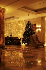 A luxurious indoor space decorated for the holiday season. A large Christmas tree adorned with lights and ornaments stands near a curtain of golden fairy lights. Elegant furniture, including armchairs and a table with a lamp, are arranged neatly. The marble flooring reflects the warm, festive lighting.