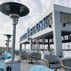 An outdoor dining area with stylish, modern furniture featuring blue and white chairs and cushions. The establishment has a sleek, contemporary design with large, prominent signage that reads 'MEZZA HOUSE'. Several patio heaters are visible, and potted plants add a touch of greenery to the space. The sky is partially cloudy, contributing to a calm atmosphere.