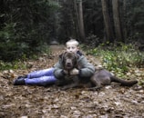A young person lies on the forest floor, embracing a large brown dog. The setting is a woodland path covered with fallen leaves, surrounded by dense trees and greenery. The atmosphere is calm and natural, with the person and dog appearing relaxed and content.