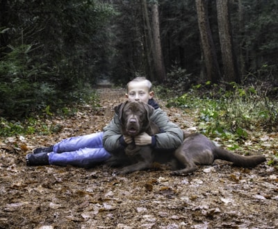 A young person lies on the forest floor, embracing a large brown dog. The setting is a woodland path covered with fallen leaves, surrounded by dense trees and greenery. The atmosphere is calm and natural, with the person and dog appearing relaxed and content.