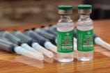 Two vials labeled 'COVISHIELD ChAdOx1 nCoV-19 Corona Virus Vaccine (Recombinant)' are positioned upright on a wooden surface. These vials are accompanied by several syringes with needles, which are laid down parallel behind the vials.
