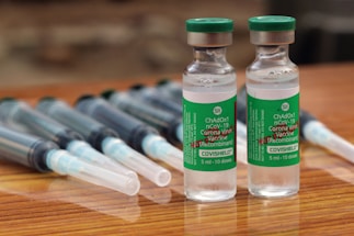 Two vials labeled 'COVISHIELD ChAdOx1 nCoV-19 Corona Virus Vaccine (Recombinant)' are positioned upright on a wooden surface. These vials are accompanied by several syringes with needles, which are laid down parallel behind the vials.
