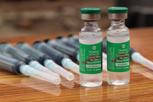 Two vials labeled 'COVISHIELD ChAdOx1 nCoV-19 Corona Virus Vaccine (Recombinant)' are positioned upright on a wooden surface. These vials are accompanied by several syringes with needles, which are laid down parallel behind the vials.