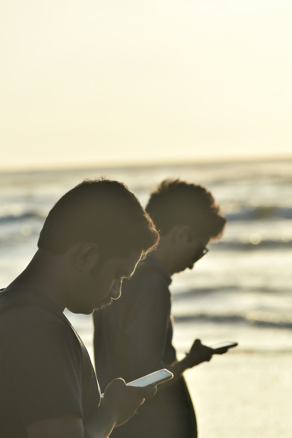 two men standing on the beach looking at their cell phones