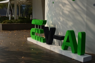 A display of large, stylized letters in green and black set against a simple, modern background. The letters are placed on a white platform on a wooden deck, surrounded by scattered autumn leaves. In the background, there is greenery and modern architectural elements.