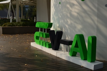 A display of large, stylized letters in green and black set against a simple, modern background. The letters are placed on a white platform on a wooden deck, surrounded by scattered autumn leaves. In the background, there is greenery and modern architectural elements.