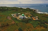 An aerial view of a mosque with a green dome surrounded by lush greenery and agricultural fields, extending towards the coastline where the blue ocean meets the land. Several small buildings and roads are scattered throughout the landscape, creating a peaceful and picturesque rural setting.