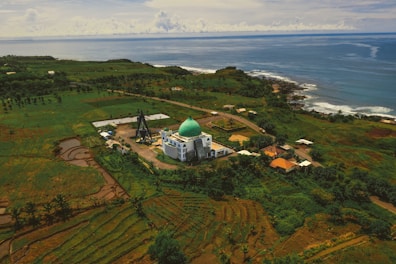 An aerial view of a mosque with a green dome surrounded by lush greenery and agricultural fields, extending towards the coastline where the blue ocean meets the land. Several small buildings and roads are scattered throughout the landscape, creating a peaceful and picturesque rural setting.