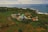 An aerial view of a mosque with a green dome surrounded by lush greenery and agricultural fields, extending towards the coastline where the blue ocean meets the land. Several small buildings and roads are scattered throughout the landscape, creating a peaceful and picturesque rural setting.