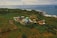 An aerial view of a mosque with a green dome surrounded by lush greenery and agricultural fields, extending towards the coastline where the blue ocean meets the land. Several small buildings and roads are scattered throughout the landscape, creating a peaceful and picturesque rural setting.