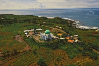 An aerial view of a mosque with a green dome surrounded by lush greenery and agricultural fields, extending towards the coastline where the blue ocean meets the land. Several small buildings and roads are scattered throughout the landscape, creating a peaceful and picturesque rural setting.