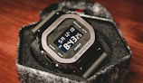 A digital wristwatch with a metallic bezel and a rugged design, displaying the time 8:15:35 and the date Saturday, 12-18. The watch is a G-Shock model by Casio, with a prominent display and robust buttons on the side. It sits in a textured box that complements its sturdy build.