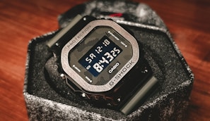 A digital wristwatch with a metallic bezel and a rugged design, displaying the time 8:15:35 and the date Saturday, 12-18. The watch is a G-Shock model by Casio, with a prominent display and robust buttons on the side. It sits in a textured box that complements its sturdy build.