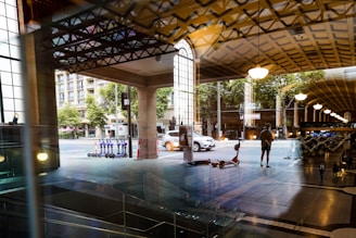 A view through a glass window reveals a blend of indoor and outdoor scenes. The interior has a patterned ceiling with hanging lights and stairs leading downwards. Outside, a street is visible with a parked white car, row of electric scooters, and trees. Pedestrians are walking, and urban architecture including building facades surrounds the area.