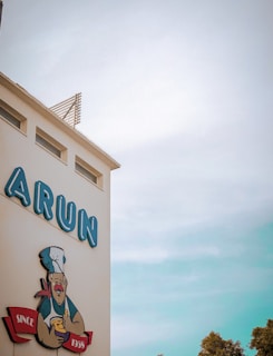 A building facade with prominently displayed signage in block letters spelling 'ARUN' and a caricature of a chef holding an ice cream cone. The chef is also shown with a red scarf, and underneath is a banner reading 'SINCE 1956'. The sky is clear with subtle blue tones, and some foliage is visible at the bottom of the image.