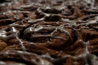 A close-up view of freshly baked pastries drizzled with icing. The image highlights the swirls of the brown pastry dough and the glossy texture of the light-colored icing.