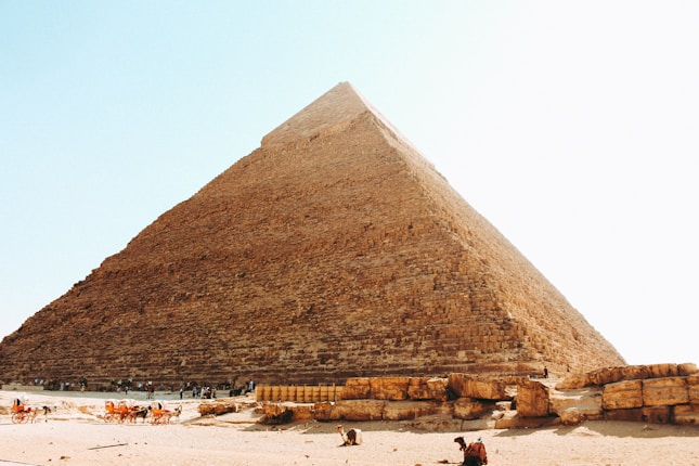 A large, ancient pyramid made of stone blocks rises majestically into a clear blue sky. At the base of the pyramid, there are tourists and camels, adding a sense of scale and human presence to the scene. The pyramid's surface shows the texture of the weathered stones, indicating its age and historical significance.