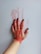 A hand with black-painted nails is pressed against a surface, leaving a smeared red paint imprint. The fingers and palm are covered with red paint, and the background is a plain, light-colored surface.