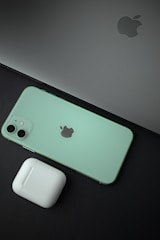 A sleek green smartphone, likely an iPhone, is placed next to a pair of white wireless earbuds in their charging case. On the upper part of the frame is a laptop with a visible logo, suggesting a cohesively branded set of electronics.