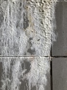 A close-up view of a textured wall with efflorescence. The surface appears rough and uneven, with white salt deposits covering sections of the gray concrete tiles.