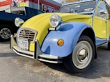 A vintage Citroën 2CV car with a two-tone yellow and blue paint scheme parked outside an auto care center. The vehicle has distinct round headlights and a classic body design, with a New York license plate on the front bumper. To the side, there is a dark-colored pickup truck, and a wall with signage and an American flag.
