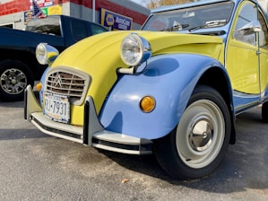 A vintage Citroën 2CV car with a two-tone yellow and blue paint scheme parked outside an auto care center. The vehicle has distinct round headlights and a classic body design, with a New York license plate on the front bumper. To the side, there is a dark-colored pickup truck, and a wall with signage and an American flag.