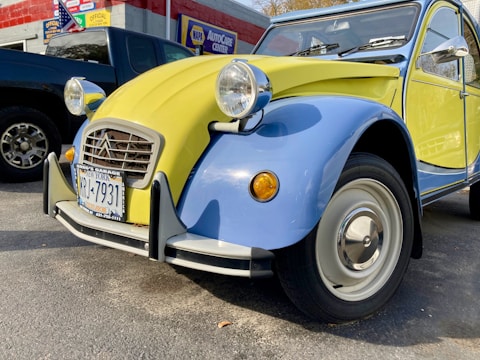 A vintage Citroën 2CV car with a two-tone yellow and blue paint scheme parked outside an auto care center. The vehicle has distinct round headlights and a classic body design, with a New York license plate on the front bumper. To the side, there is a dark-colored pickup truck, and a wall with signage and an American flag.