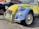 A vintage Citroën 2CV car with a two-tone yellow and blue paint scheme parked outside an auto care center. The vehicle has distinct round headlights and a classic body design, with a New York license plate on the front bumper. To the side, there is a dark-colored pickup truck, and a wall with signage and an American flag.