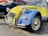 A vintage Citroën 2CV car with a two-tone yellow and blue paint scheme parked outside an auto care center. The vehicle has distinct round headlights and a classic body design, with a New York license plate on the front bumper. To the side, there is a dark-colored pickup truck, and a wall with signage and an American flag.