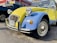 A vintage Citroën 2CV car with a two-tone yellow and blue paint scheme parked outside an auto care center. The vehicle has distinct round headlights and a classic body design, with a New York license plate on the front bumper. To the side, there is a dark-colored pickup truck, and a wall with signage and an American flag.