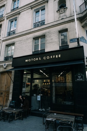 A small coffee shop named Motors Coffee is situated on a street corner in an urban area. The building is tall with multiple windows and an ornate architectural design. Outside, a couple of wooden tables and chairs are placed, with two people seated, possibly having a conversation. The street signs read 'Rue des Halles', indicating a potential location in a French-speaking area.