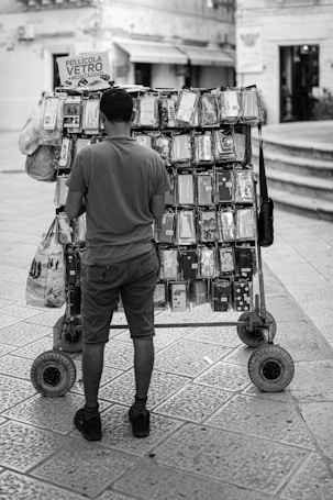 A person stands in front of a mobile street vendor cart displaying various smartphone cases. The cart is parked on a cobblestone street, and the cases are organized neatly in rows. Signs on the cart suggest the sale includes screen protectors with installation services. The background shows a building, steps, and a storefront.