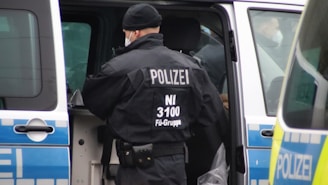 A police officer, wearing a black uniform with 'POLIZEI' written on the back, stands next to a police van with its door open. The vehicle has blue and white markings. The officer is wearing a cap and a mask.