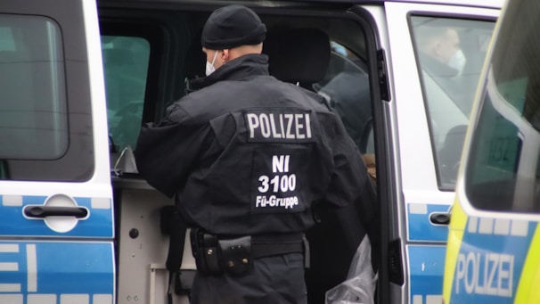 A police officer, wearing a black uniform with 'POLIZEI' written on the back, stands next to a police van with its door open. The vehicle has blue and white markings. The officer is wearing a cap and a mask.