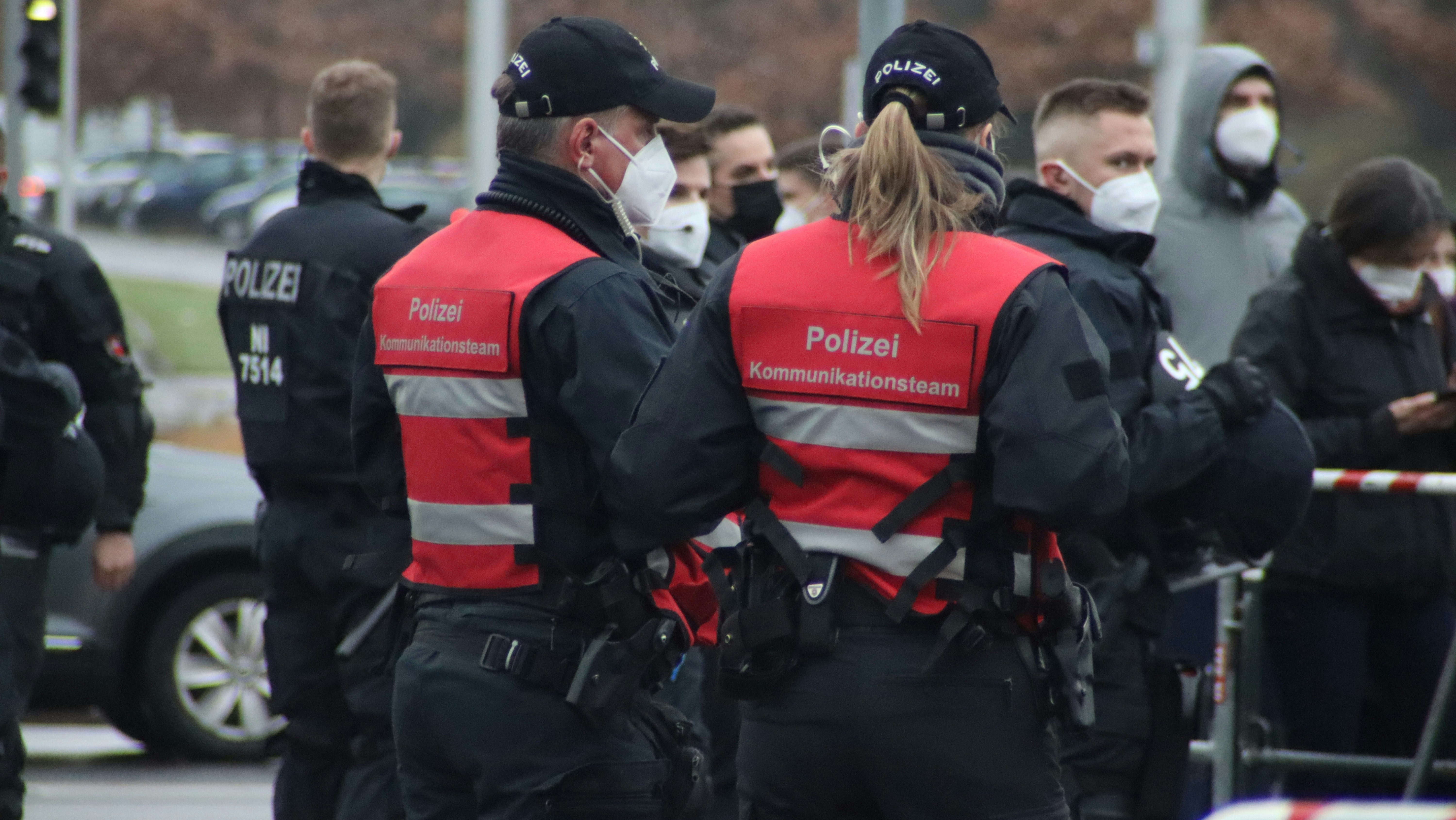 A group of uniformed police officers wearing red vests marked 'Kommunikationsteam' are interacting with people while wearing face masks. The scene seems to be part of a larger public gathering or event. Several officers and individuals in the background are also seen wearing protective masks.