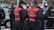 A group of uniformed police officers wearing red vests marked 'Kommunikationsteam' are interacting with people while wearing face masks. The scene seems to be part of a larger public gathering or event. Several officers and individuals in the background are also seen wearing protective masks.