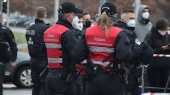 A group of uniformed police officers wearing red vests marked 'Kommunikationsteam' are interacting with people while wearing face masks. The scene seems to be part of a larger public gathering or event. Several officers and individuals in the background are also seen wearing protective masks.