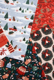 A collection of festive wrapping papers featuring winter and holiday themes. One pattern shows snowy landscapes with dogs, presents, trees, and a red truck. Another design has red and green holly with bows, while another showcases Christmas wreaths tied with red ribbons. A final pattern includes mittens, sweaters, and various ornaments on a dark green background.