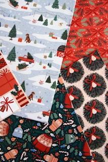 A collection of festive wrapping papers featuring winter and holiday themes. One pattern shows snowy landscapes with dogs, presents, trees, and a red truck. Another design has red and green holly with bows, while another showcases Christmas wreaths tied with red ribbons. A final pattern includes mittens, sweaters, and various ornaments on a dark green background.
