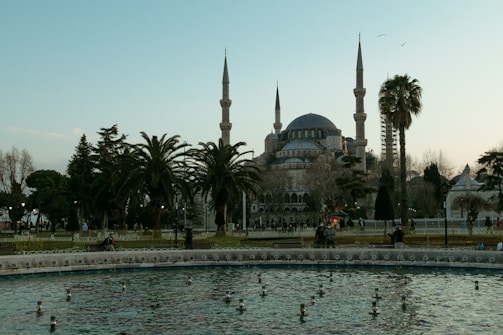 An iconic historical mosque with multiple minarets stands prominently against the sky. Surrounding it are trees, including tall palm trees, and a large water fountain in the foreground. A few people are scattered around the area, enjoying the serene atmosphere.