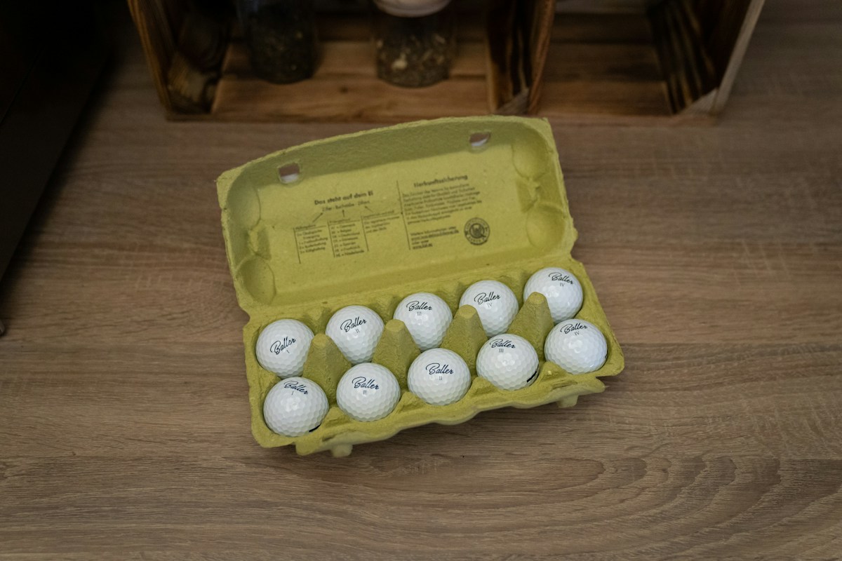 A carton of twelve white golf balls on a wooden floor, resembling Easter eggs for creative Easter egg hunt ideas.