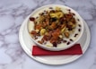 A bowl of cereal is topped with a variety of dried fruits and almonds, served with milk. The bowl is placed on a white plate with a red napkin under the bowl. The background is a marble-like white surface with grey veins.
