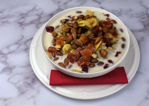 A bowl of cereal is topped with a variety of dried fruits and almonds, served with milk. The bowl is placed on a white plate with a red napkin under the bowl. The background is a marble-like white surface with grey veins.