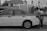 A black and white photo shows a sedan with visible damage and tape on the rear door. Two individuals are standing at the open trunk, engaged in arranging or examining its contents. The background features a building with a tiled roof and exposed piping.